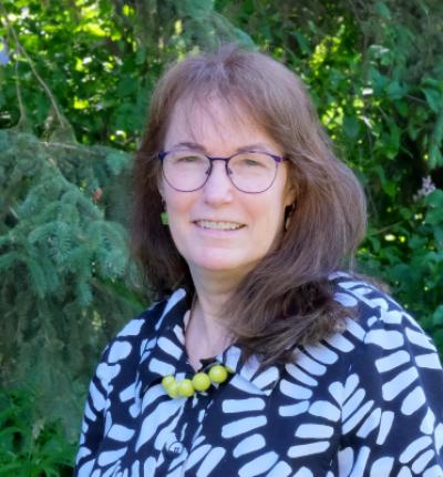 A woman with shoulder length hair and glasses is smiling and standing in front of evergreen trees.