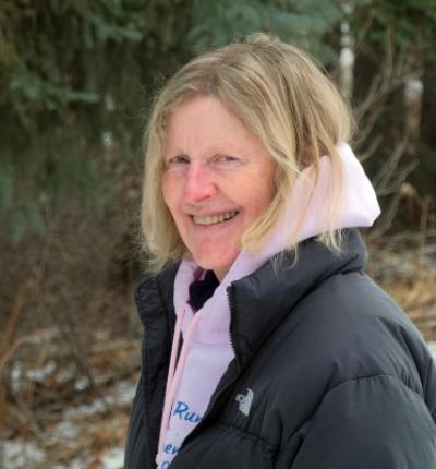 A woman with neck length blonde hair wearing a winter jacket standing in front of a blurry evergreen forest background.