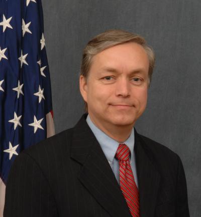 A man in a dark suit with a red tie standing just in front of a United States flag pole