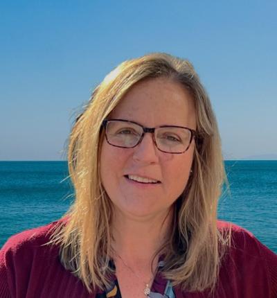 Dr. Jill Lewandowski with shoulder length hair is wearing glasses and posing for a portrait in front of an ocean background.