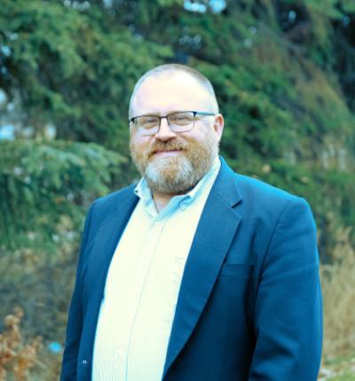 Portrait of Givey Kochanowski wearing office casual clothes in front of evergreen trees.