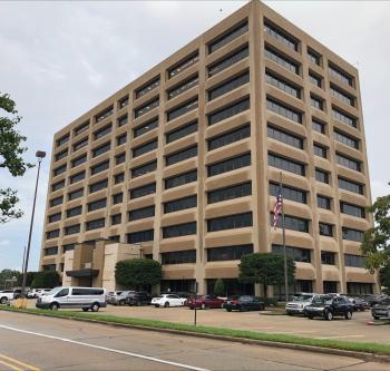 A 10-story tall brutalist concrete building surrounded by a mostly full parking lot.