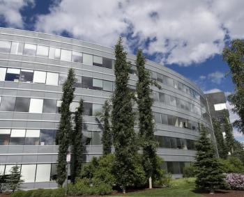 Looking up from the ground, a 5-story tall, semi-arced building with all evergreen trees in the foreground.