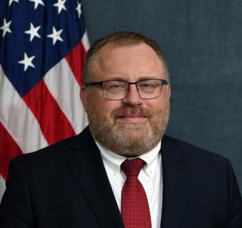 Headshot portrait of Givey Kochanowski wearing a dark suit, red tie, short cropped hair and a neatly trimmed beard.