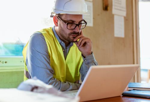 A person wearing a white hardhat, a yellow safety vest sitting in front of an open laptop.