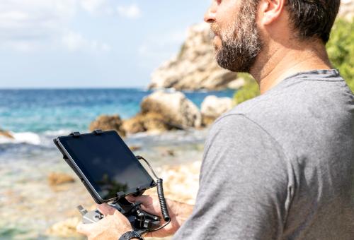 A person standing near an ocean cove holding a tablet PC in their hands.